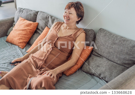 Unaltered senior woman relaxing at home and watching a comedy on TV, enjoying a cozy evening in her living room. Concept of leisure, comfort, positive emotions, mental wellbeing and everyday lifestyle Unaltered senior woman relaxing at home and watching a comedy on TV, enjoying a cozy evening in her living room. Concept of leisure, comfort, positive emotions, mental wellbeing and everyday lifestyle 133668628