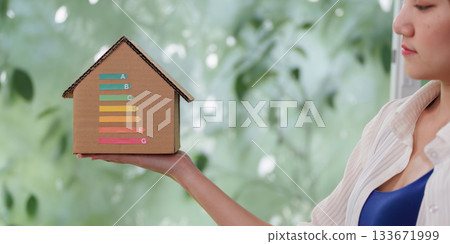 Energy Efficiency. Woman holding a cardboard house model with energy rating labels. Energy Efficiency. Woman holding a cardboard house model with energy rating labels. 133671999