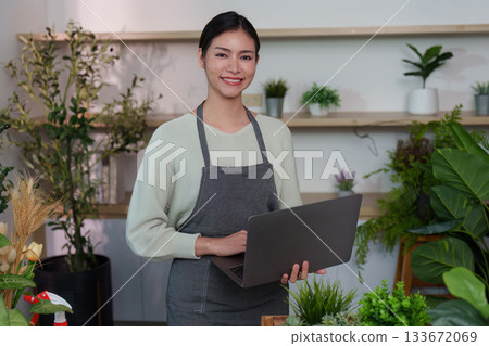 Indoor Gardening. Smiling woman using a laptop in a plant-filled workspace. Indoor Gardening. Smiling woman using a laptop in a plant-filled workspace. 133672069