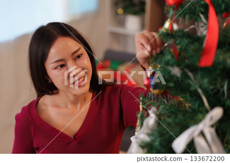 SSTK Christmas. Woman smiling while hanging a colorful ornament on a Christmas tree. SSTK Christmas. Woman smiling while hanging a colorful ornament on a Christmas tree. 133672200