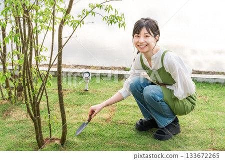 一位年輕的亞裔女子身穿圍裙，手持鐵鍬，正在種植園裡的樹木。 133672265