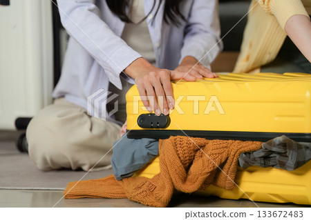 Travel Planning. Friends struggling to close an overstuffed yellow suitcase for their trip. 133672483
