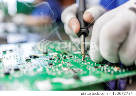 Closeup of worker in protective gloves soldering electronic circuit board with hot iron and visible smoke during assembly process 133672784