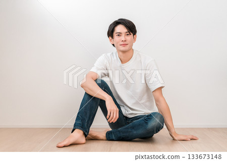 一位身穿牛仔褲的年輕亞裔男子坐在白牆的客廳裡 一位身穿牛仔褲的年輕亞裔男子坐在白牆的客廳裡 133673148