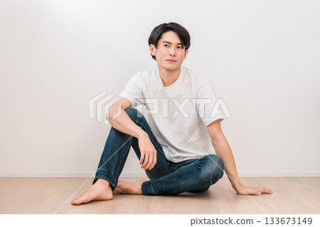 一位身穿牛仔褲的年輕亞裔男子坐在白牆的客廳裡 一位身穿牛仔褲的年輕亞裔男子坐在白牆的客廳裡 133673149