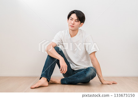 一位身穿牛仔褲的年輕亞裔男子坐在白牆的客廳裡 133673150