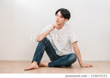 一位身穿牛仔褲的年輕亞裔男子坐在白牆的客廳裡 一位身穿牛仔褲的年輕亞裔男子坐在白牆的客廳裡 133673151