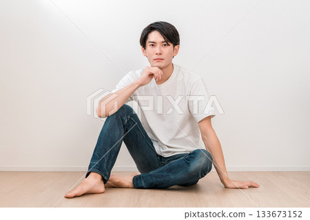 A young Asian man in jeans sitting in a living room with white walls 133673152