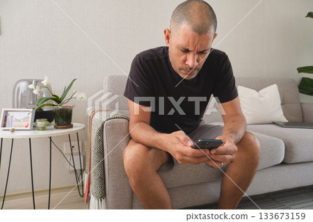 Man feeling concerned looking at smartphone on couch 133673159