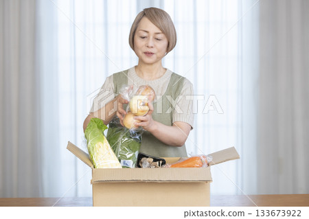 A senior woman rejoices in the delivery of vegetables delivered directly from the farm in her living room A senior woman rejoices in the delivery of vegetables delivered directly from the farm in her living room 133673922