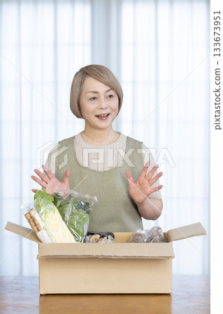 A senior woman rejoices in the delivery of vegetables delivered directly from the farm in her living room A senior woman rejoices in the delivery of vegetables delivered directly from the farm in her living room 133673951