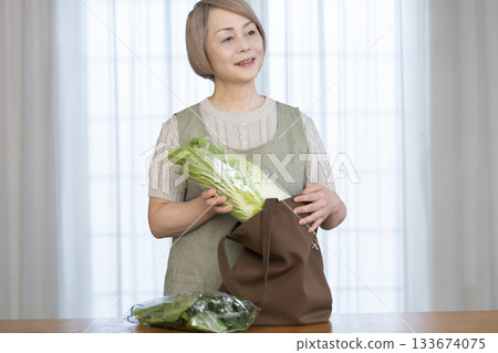 Senior woman taking vegetables out of a shopping bag in the living room Senior woman taking vegetables out of a shopping bag in the living room 133674075