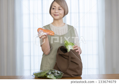 Senior woman taking vegetables out of a shopping bag in the living room Senior woman taking vegetables out of a shopping bag in the living room 133674079