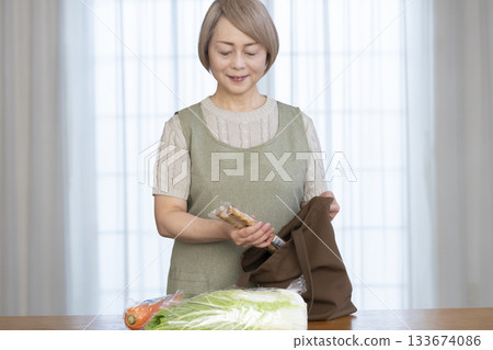 Senior woman taking vegetables out of a shopping bag in the living room Senior woman taking vegetables out of a shopping bag in the living room 133674086