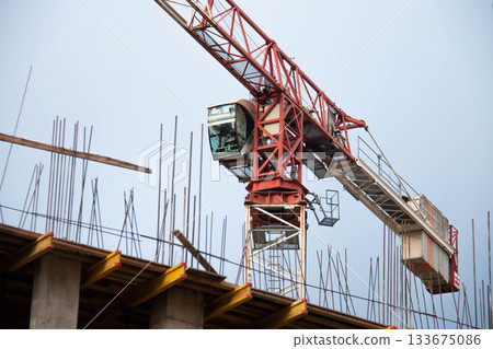 Close-up of Control Cabin of Red Tower Crane with Counterweight at Construction Site. In Foreground are Yellow Formwork Beams with Monolithic Box Fittings. Close-up of Control Cabin of Red Tower Crane with Counterweight at Construction Site. In Foreground are Yellow Formwork Beams with Monolithic Box Fittings. 133675086