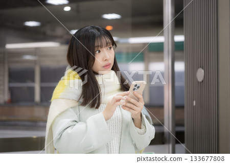 A woman wearing a cute scarf and holding a smartphone in what looks like a winter station platform/bus terminal. Is she delayed? 133677808