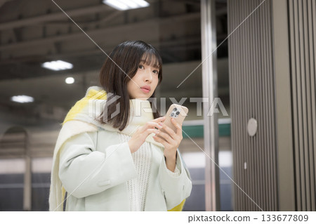 A woman wearing a cute scarf and holding a smartphone in what looks like a winter station platform/bus terminal. Is she delayed? 133677809