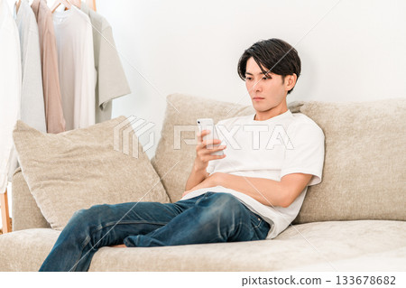 A young Asian man sitting on the living room sofa and looking at his smartphone (lazy, lazy) A young Asian man sitting on the living room sofa and looking at his smartphone (lazy, lazy) 133678682