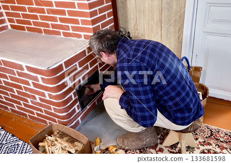Mature man lights wood stove in country house. 133681598