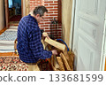 Preparing firewood for brick stove, middle aged man seated on low stool lifts log from bark basket to place into firebox before kindling burn. 133681599