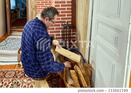 Preparing firewood for brick stove, middle aged man seated on low stool lifts log from bark basket to place into firebox before kindling burn. 133681599