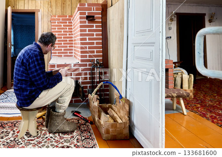 Mature man sits on low stool preparing kindling before brick stove. Mature man sits on low stool preparing kindling before brick stove. 133681600