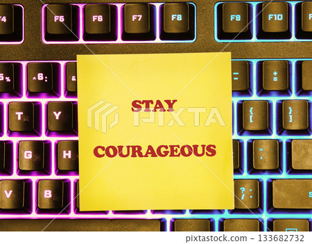 A vibrant keyboard shines with colorful lights, featuring a yellow sticky note that reads Stay Courageous, adding inspiration to any creative workspace or study area 133682732