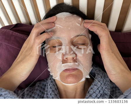 Mature woman applying a moisturizing sheet facial mask at home. Anti aging skincare routine for mature skin, hydration, rejuvenation, fine lines treatment, beauty self care concept 133683136