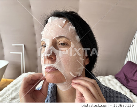 Mature woman applying a moisturizing sheet facial mask at home. Anti aging skincare routine for mature skin, hydration, rejuvenation, fine lines treatment, beauty self care concept 133683139