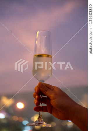 A hand holding a glass of Prosecco or sparkling wine with colorful sunset sky and city view on the background A hand holding a glass of Prosecco or sparkling wine with colorful sunset sky and city view on the background 133683220