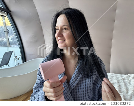 Woman Drying Hair With Compact Pink Hair Dryer at Home, Daily Beauty Routine in Cozy Bathrobe Woman Drying Hair With Compact Pink Hair Dryer at Home, Daily Beauty Routine in Cozy Bathrobe 133683272