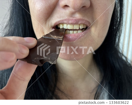 Woman sitting indoors biting a piece of dark chocolate, relaxed casual moment enjoying a sweet treat in a cozy home environment 133683622
