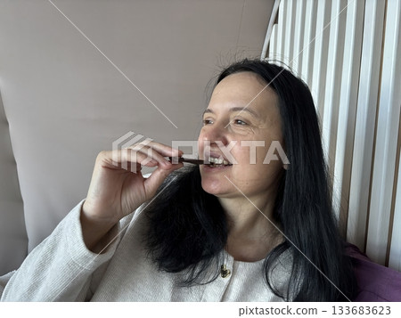 Woman sitting indoors biting a piece of dark chocolate, relaxed casual moment enjoying a sweet treat in a cozy home environment 133683623