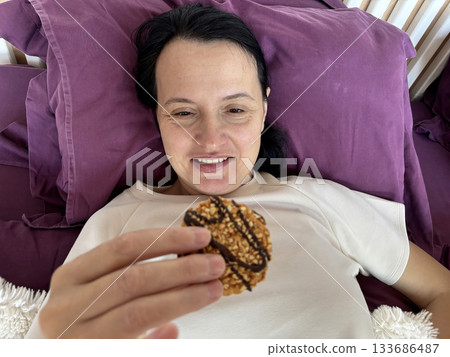 Smiling adult woman eating, tasting cookies while resting on the bed, cozy breakfast or snack moment in a bright peaceful bedroom Smiling adult woman eating, tasting cookies while resting on the bed, cozy breakfast or snack moment in a bright peaceful bedroom 133686487