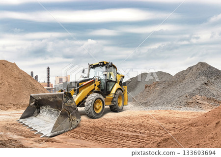 A yellow backhoe loader moves dirt and gravel at a construction site. Heavy clouds fill the sky, hinting at possible rain in the area A yellow backhoe loader moves dirt and gravel at a construction site. Heavy clouds fill the sky, hinting at possible rain in the area 133693694