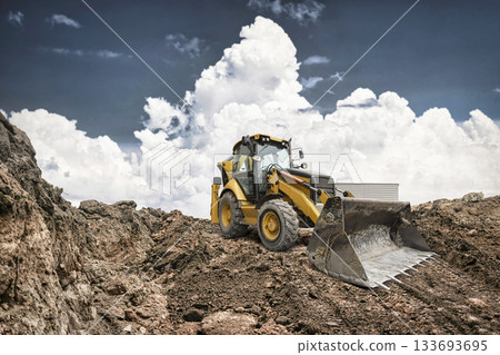 A construction vehicle with a large front bucket works on a dirt site, moving earth and rocks under a bright, cloudy sky A construction vehicle with a large front bucket works on a dirt site, moving earth and rocks under a bright, cloudy sky 133693695