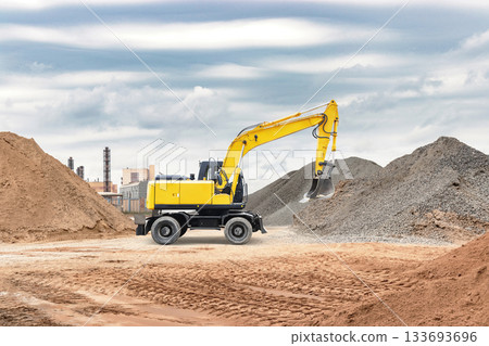 A yellow excavator operates on a construction site, digging and moving material next to large mounds of sand and gravel under an overcast sky A yellow excavator operates on a construction site, digging and moving material next to large mounds of sand and gravel under an overcast sky 133693696