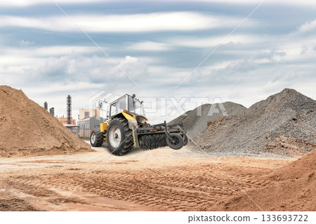 A large tractor works on flattening dirt at an industrial construction site. Nearby, there are mounds of sand and gravel under a cloudy sky 133693722