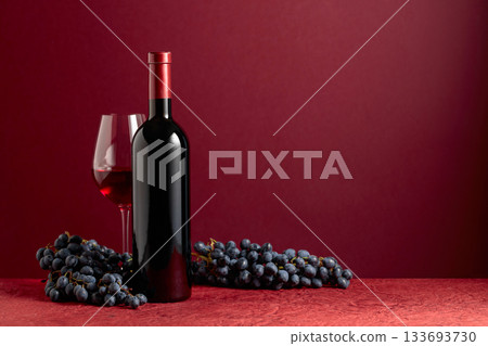 Bottle and glass of red wine with blue grapes on a red background. Bottle and glass of red wine with blue grapes on a red background. 133693730