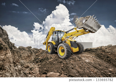 A yellow backhoe loader transports dirt at a construction site, highlighting the machinery's power against a backdrop of dramatic clouds 133693782