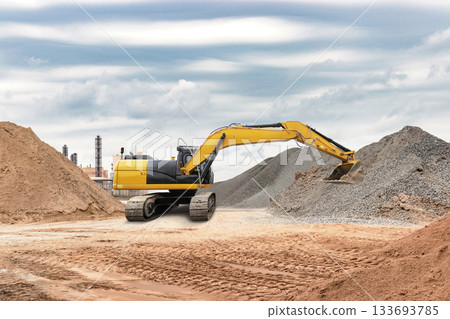 Excavator works on a construction site, moving piles of gravel around while clouds linger in the sky, indicating a busy work environment 133693785