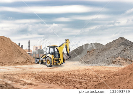 A yellow backhoe works on a construction site, digging through mounds of dirt and gravel with industrial buildings in the background on a cloudy day 133693789