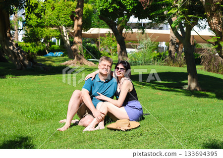 Adult friends, a man and a woman, are sitting on the green grass of a city park 133694395