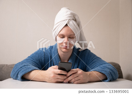 Young caucasian man wearing towel on his head and under-eye patches while using his smartphone during home skincare and relaxation routine. Mens Home Self-Care Routine 133695001