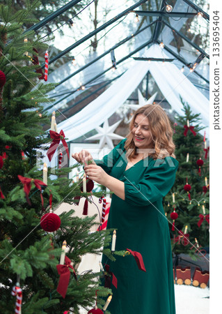 Woman decorating Christmas tree with festive ornaments in cozy winter greenhouse Woman decorating Christmas tree with festive ornaments in cozy winter greenhouse 133695404