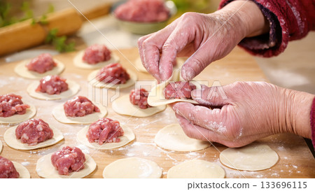 Senior hands making homemade dumplings with raw minced meat and dough on wooden table in kitchen. Traditional cooking process, close-up view. 133696115