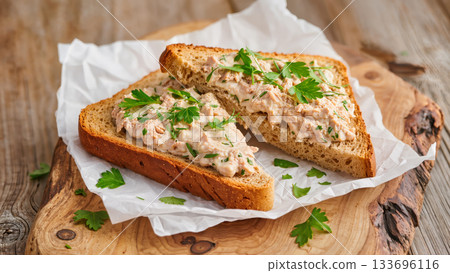 Two slices of whole grain bread with creamy tuna spread and fresh herbs on wooden board. Homemade open sandwich snack. Rustic food style. 133696116