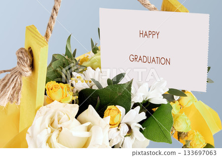 A beautiful bouquet filled with white and yellow flowers hangs against a soft blue background. A card proudly displays the words Happy Graduation, perfect for celebrating this special day 133697063
