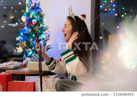 Young woman with long hair making a video call near Christmas tree and gifts in cozy festive room Young woman with long hair making a video call near Christmas tree and gifts in cozy festive room 133698495