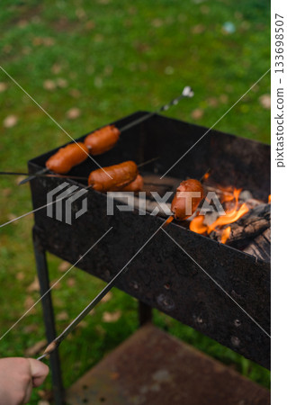 Grilling sausages on skewers over open flame in metal barbecue grill during outdoor cookout Grilling sausages on skewers over open flame in metal barbecue grill during outdoor cookout 133698507